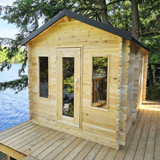 Wooden cabin with a black roof on a wooden deck by a body of water.